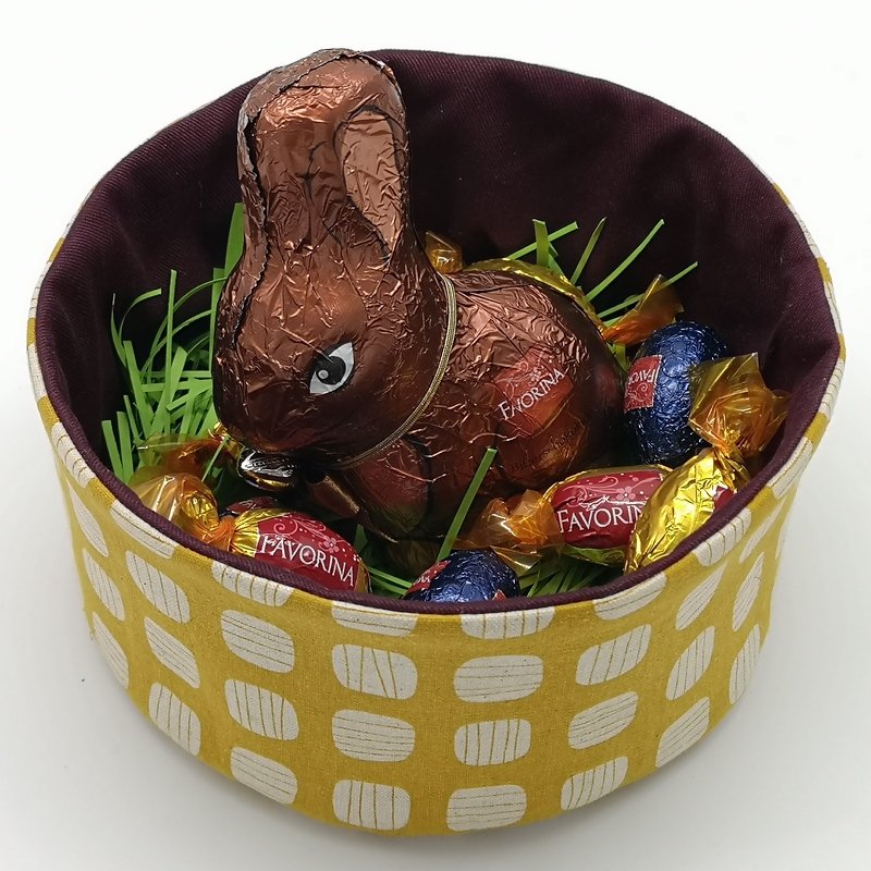 Rundes gelbes Stoff-Utensilo mit weißem Muster und dunklem Innenfutter, gefüllt mit Ostergras, Schokohase und Süßigkeiten.