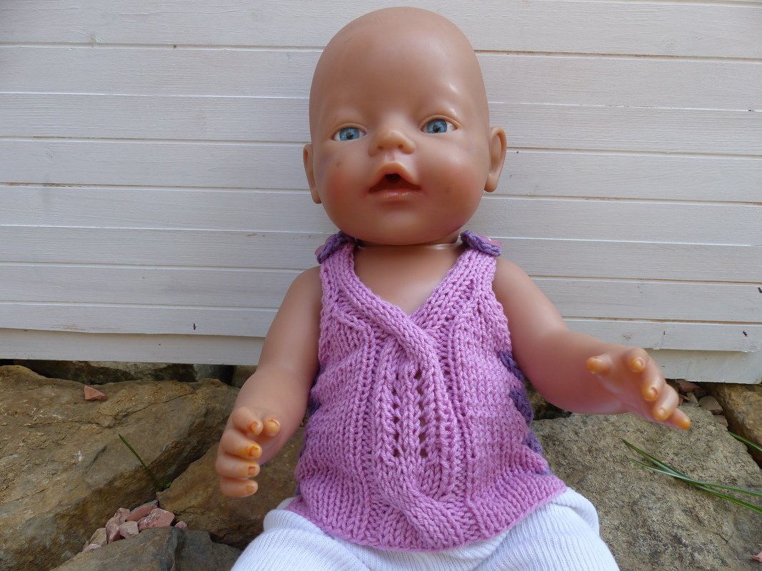 Babypuppe mit blauen Augen trägt fliederfarbenes gestricktes Top und weiße Hose, sitzt vor weißer Wand auf Steinen.