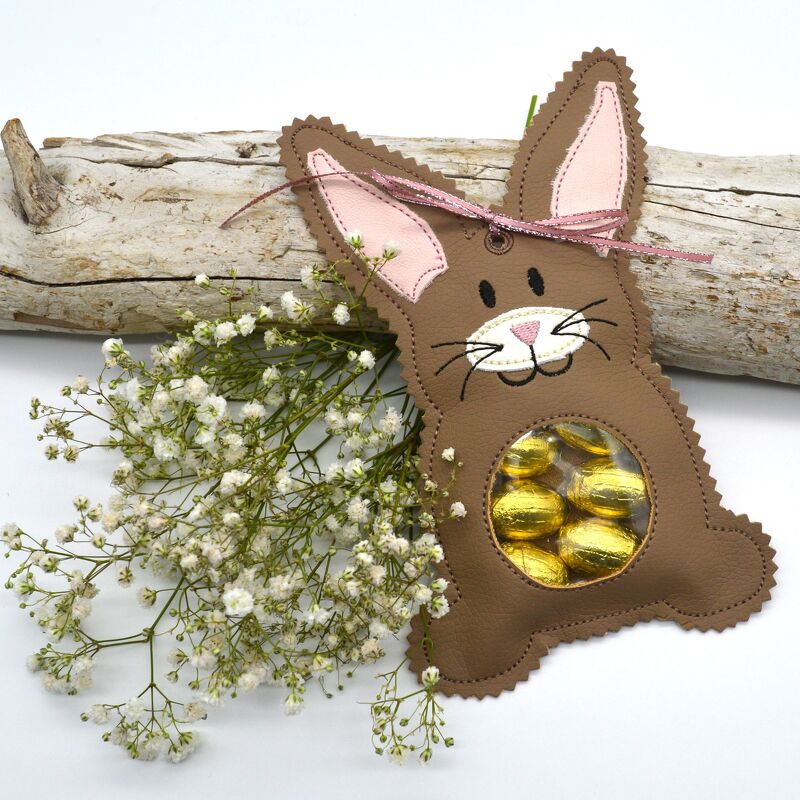 Braunes Hasentaschchen mit rosa Ohren, Schleife und rundem Sichtfenster, gefüllt mit goldenen Schokoladeneiern, dekorativ neben weißen Blüten