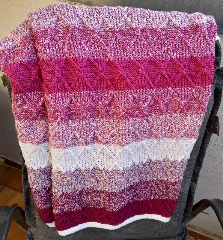 Gestrickte Babydecke mit Rautenmuster in Rosa-, Pink- und Beeretönen, über eine Rückenlehne gelegt.