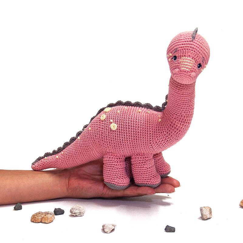 Pink crocheted long-neck dinosaur amigurumi with grey ridges, held in a hand.