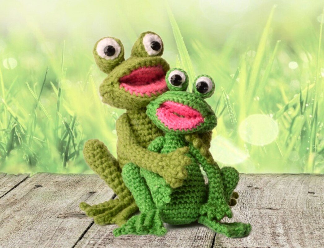 Two green crocheted amigurumi frogs, one larger holding a smaller, on a wooden surface with a blurred grassy background.