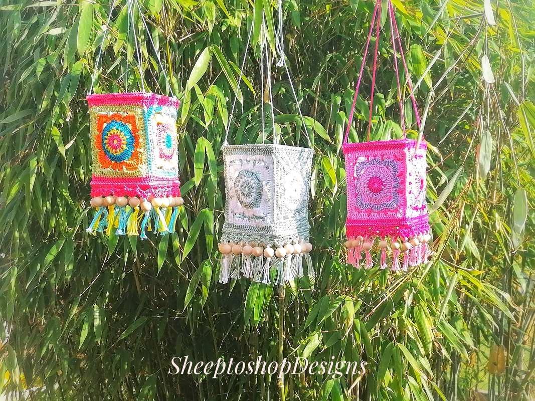 Three square crocheted lanterns in bright multicolor, pale green and pink, decorated with tassels and wooden beads, hanging against bamboo foliage.