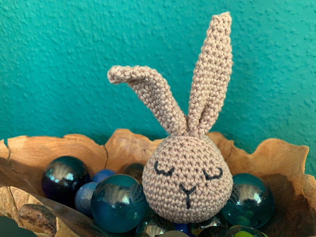 Beiger gehäkelter Amigurumi-Hase mit geschlossenen Augen in einer Holzschale voller blauer und grüner Murmeln vor türkisfarbenem Hintergrund.