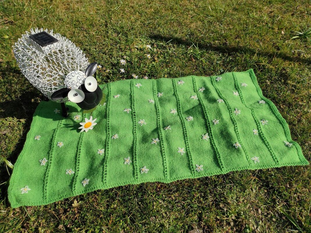 Gestrickter grüner Läufer mit weißen gestickten Blüten und Rippenstruktur, liegend auf Gras neben einer Schaffigur