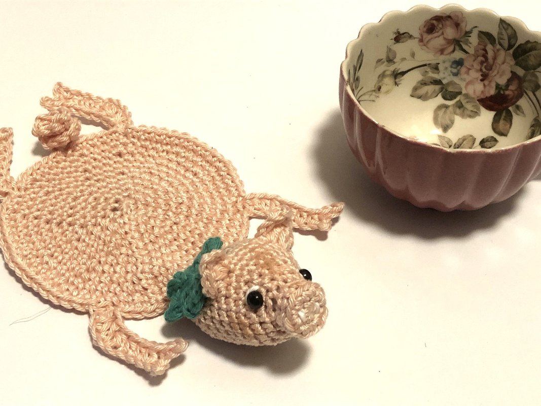 Runder gehäkelter Untersetzer in Pastellrosa mit aufgenähtem Schweinekopf und grünem Blatt, neben einer rosa Teetasse mit Blumenmuster.