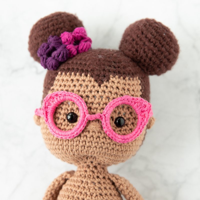 Nahaufnahme einer gehäkelten Amigurumi-Puppe mit rosa Brille, schwarzen Augen und zwei Haarknoten