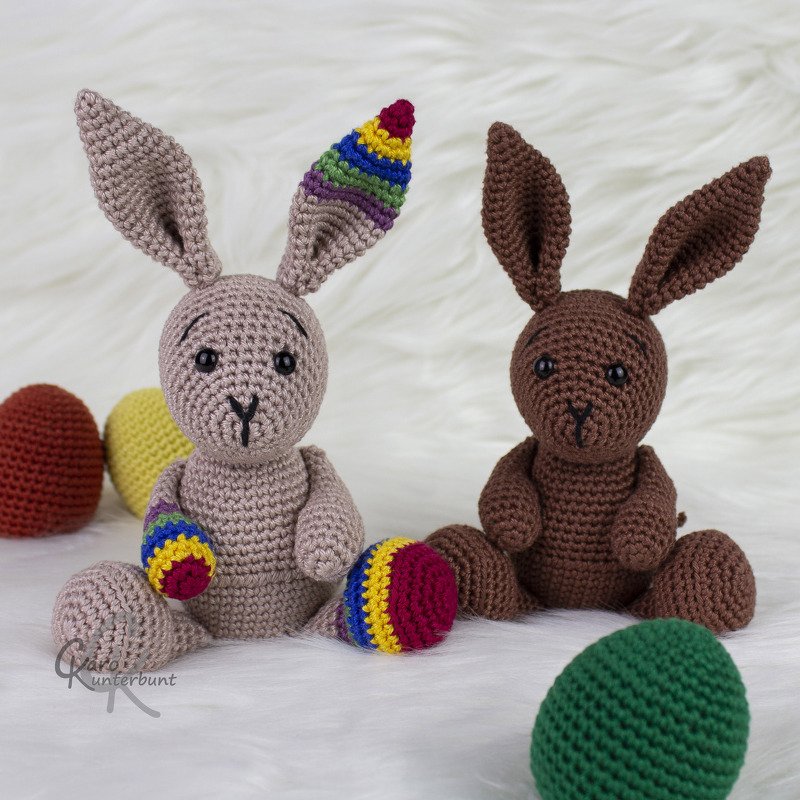 Zwei gehäkelte Amigurumi-Hasen in Beige und Braun mit bunten Streifen, umgeben von Häkel-Eiern auf hellem, flauschigem Untergrund