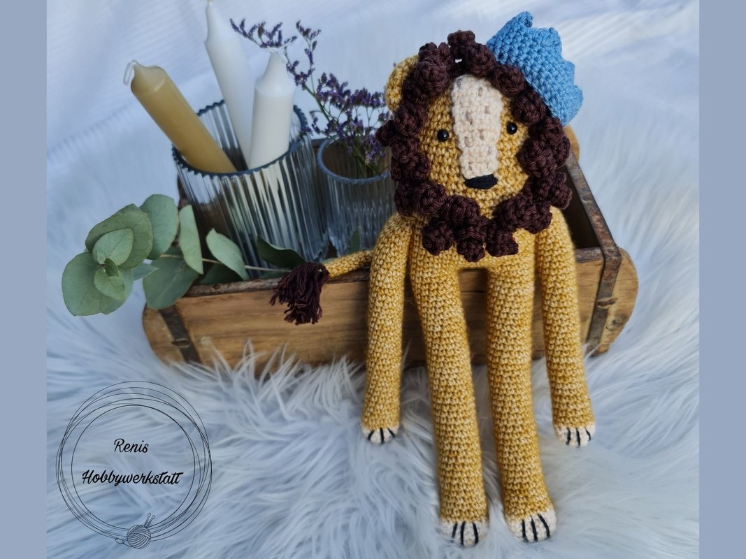 Gehäkelter Löwe mit gelbem Körper, brauner Mähne und blauer Krone auf weißem, flauschigem Untergrund.