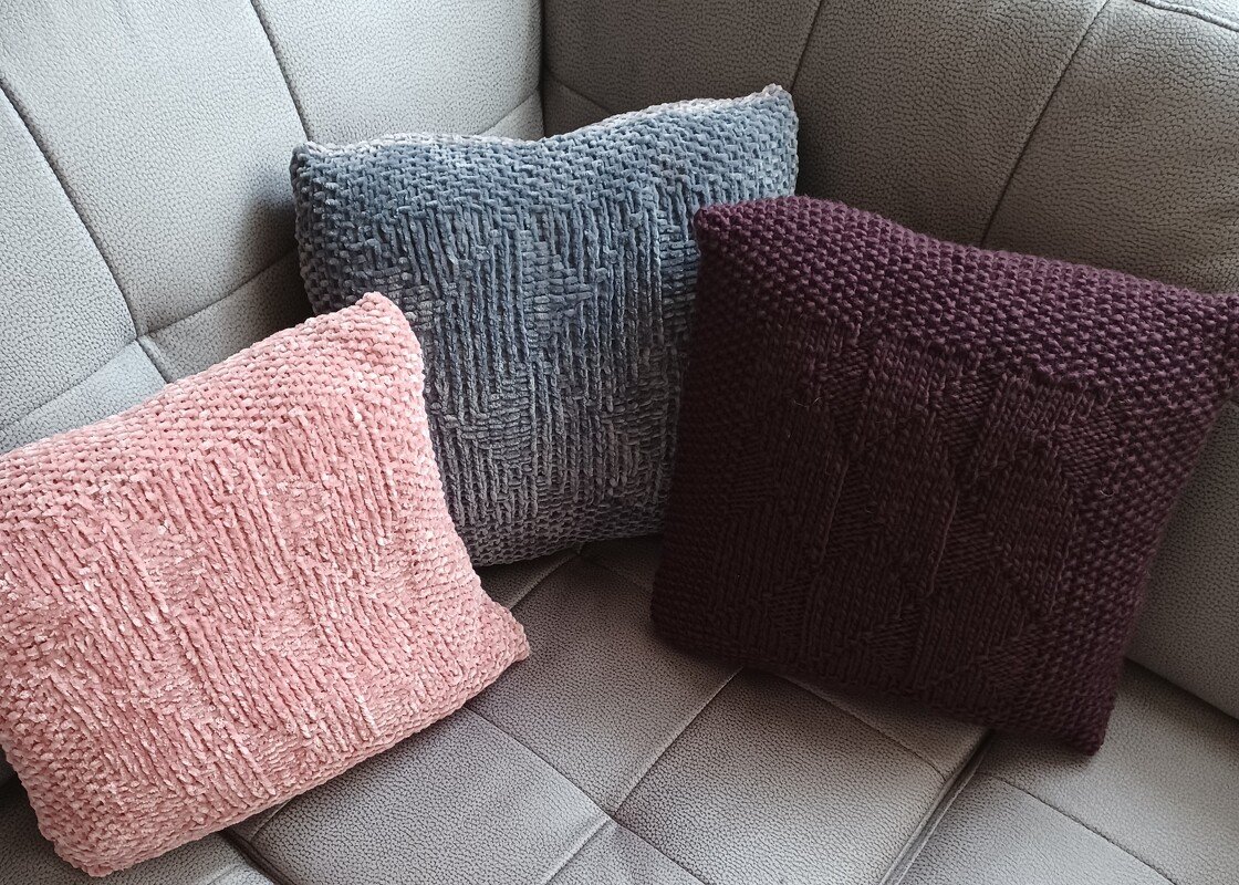 Drei gestrickte Kissen in Altrosa, Graublau und Bordeaux mit strukturierten Mustern auf einem Sofa