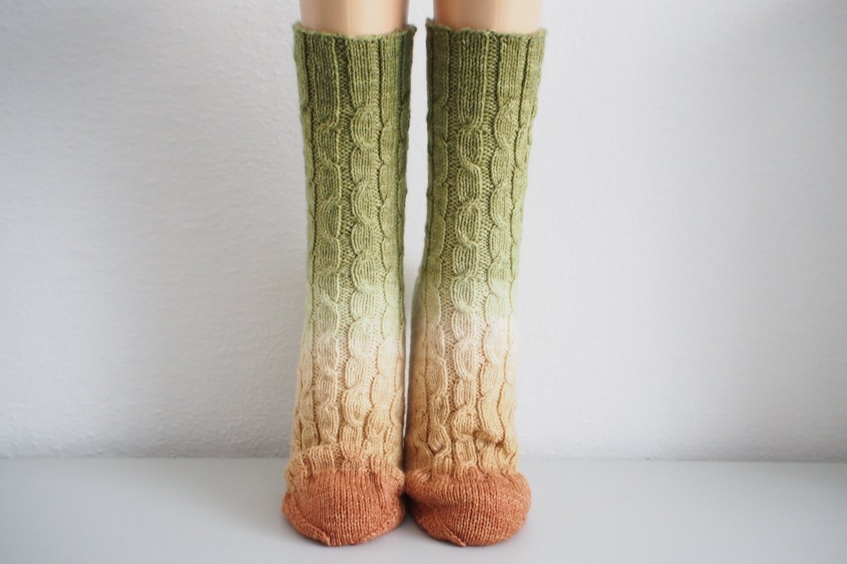 Long cable-knit socks with an ombre gradient from green to cream to rust brown and a cable pattern.
