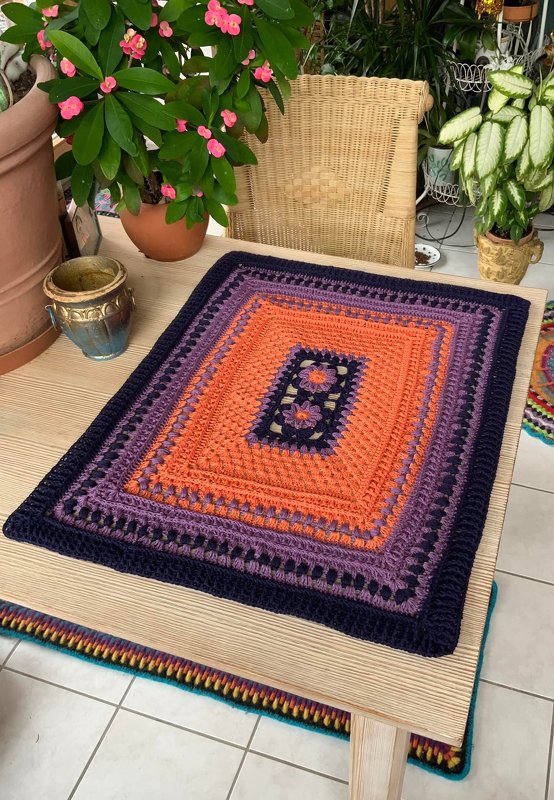 Gehäkeltes rechteckiges Deckchen mit orangefarbenem Zentrum, lila und dunkelblauem Rand und floralem Mittelmotiv auf einem Holztisch