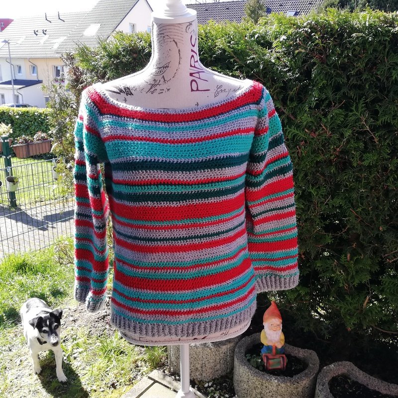 Gehäkelter Damenpullover mit roten, türkisen, grünen und grauen Querstreifen auf einer Schneiderbüste im Garten