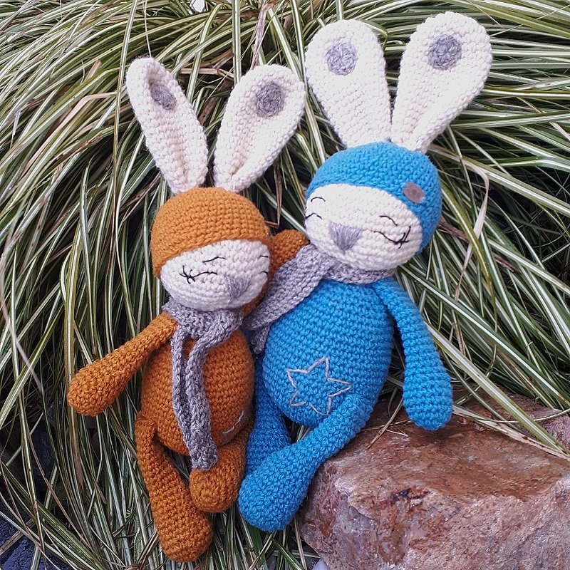 Zwei gehäkelte Amigurumi-Hasen, einer blau mit Stern auf dem Bauch, einer orange, beide mit langen Ohren und grauen Schals