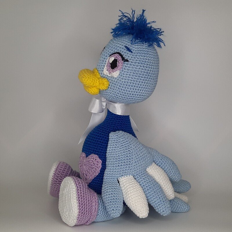 Gehäkeltes Amigurumi-Vögelchen in Hellblau und Dunkelblau mit gelbem Schnabel, großem Auge, weißer Schleife und lila Herz, sitzend vor hellem Hintergrund