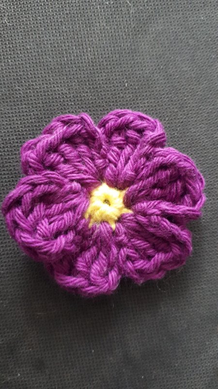 Gehäkelte Blume in Lila mit gelber Mitte auf dunklem Hintergrund