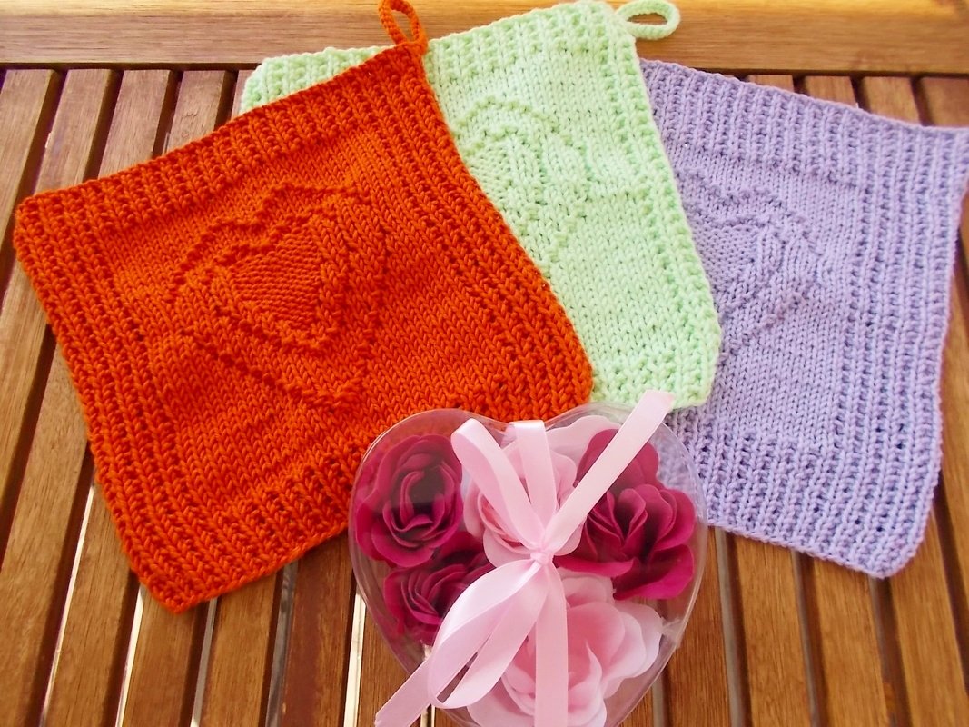 Drei gestrickte Topflappen (orange, hellgrün, flieder) mit Herzmotiv und Aufhängeschlaufen auf Holzuntergrund; herzförmige Geschenkbox mit Rosen daneben.