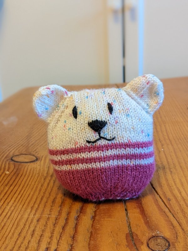 Gestricktes Amigurumi-Bärchen in Cremefarben und Magenta mit gesticktem Gesicht auf Holztisch