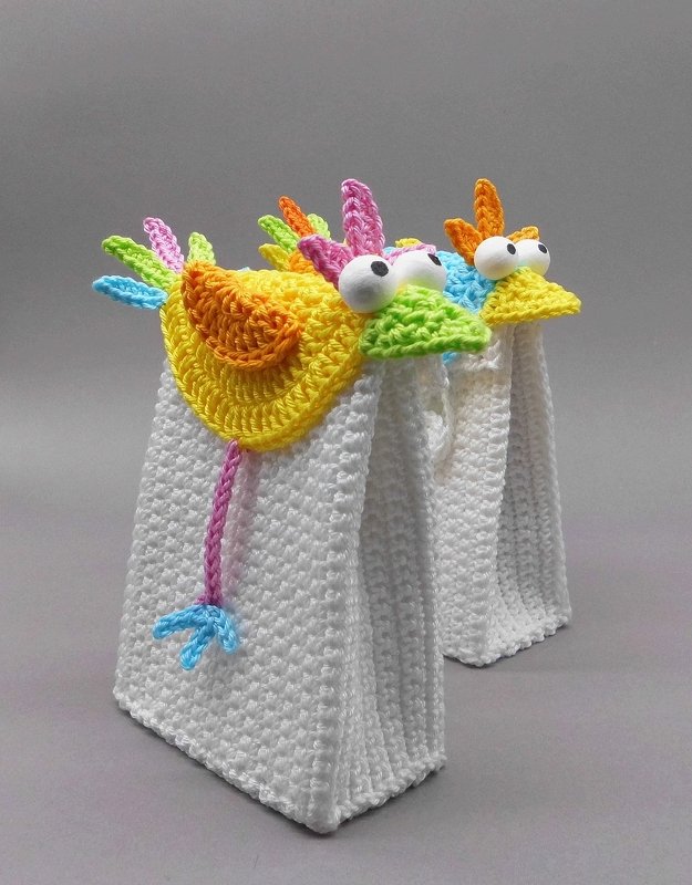 Two white crocheted rectangular gift bags with yellow bird-shaped crochet toppers, large white eyes and multicolored feather details.