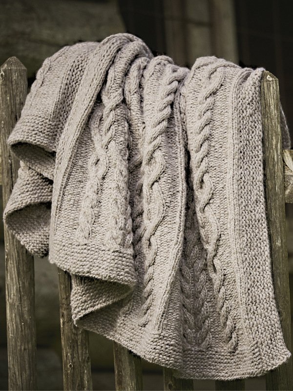 Graue Strickdecke mit Zopfmuster und kraus-rechten Maschen, über einem rustikalen Holzzaun hängend.