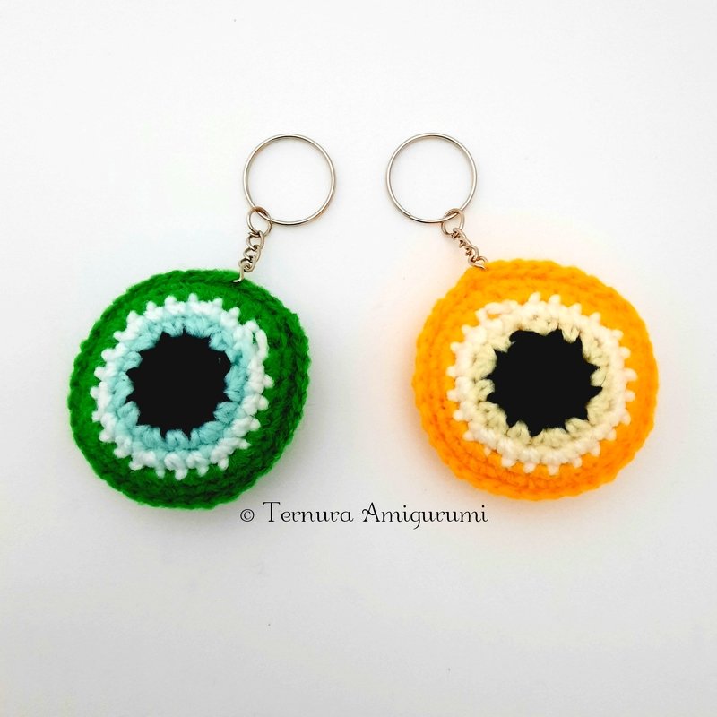 Two crocheted round keychains with concentric eye motifs, one green and one orange, both attached to metal keyrings.