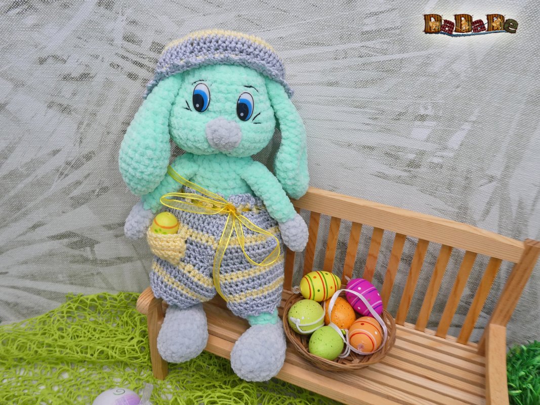Gehäkelter Amigurumi-Hase mit langer Ohren, grauer Mütze, gestreifter Hose und gelber Schleife auf einer Holzbank neben einem Körbchen mit bunten Ostereiern