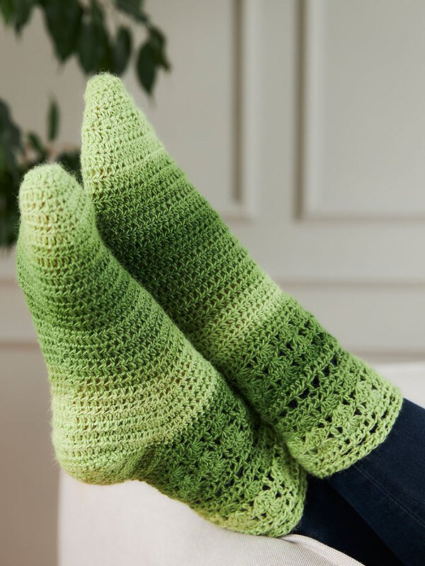 Füße mit langen gehäkelten Socken in Grüntönen mit sanftem Farbverlauf auf hellem Untergrund