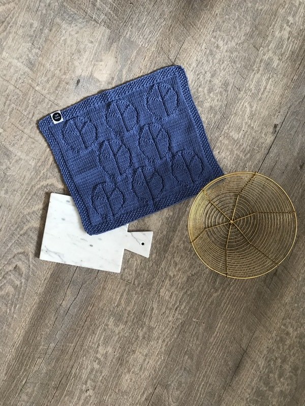 Blauer gestrickter Topflappen mit Tulpenmuster auf Holzfußboden neben Marmorbrett und Drahtkorb