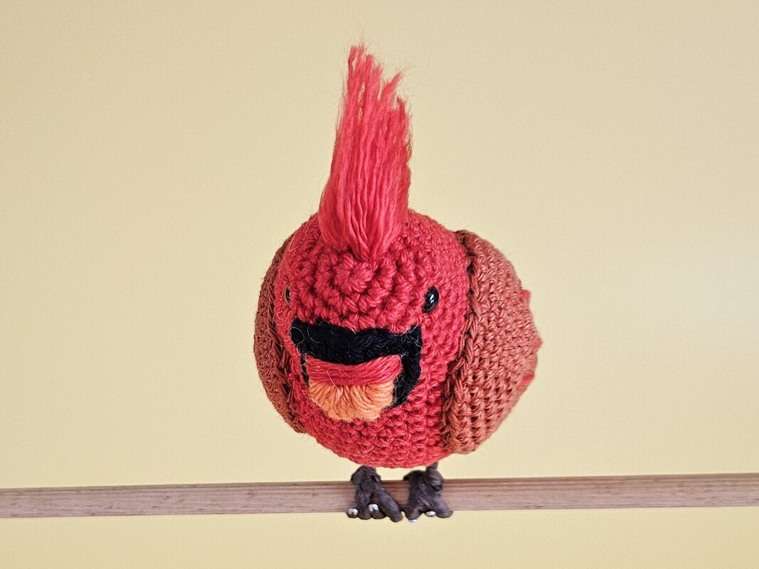 Roter gehäkelter Amigurumi‑Vogel mit aufgestellter Haube auf Holzstange