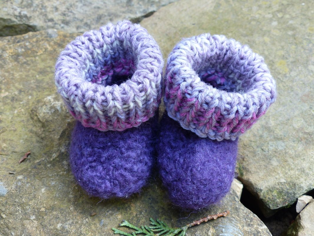 Lila gestrickte Babyschuhe mit melierten Umschlagbündchen auf Steinen