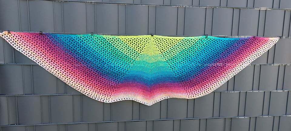 Halbrundes gehäkeltes Tuch mit buntem Farbverlauf an einem Zaun hängend