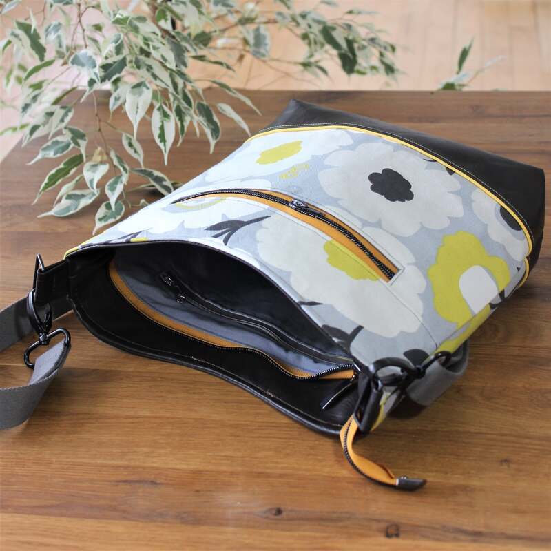 Schultertasche mit floralem Muster in Grau, Gelb und Schwarz, Kunstlederboden, orangefarbene Reißverschlüsse und verstellbarer Riemen auf Holzuntergrund.