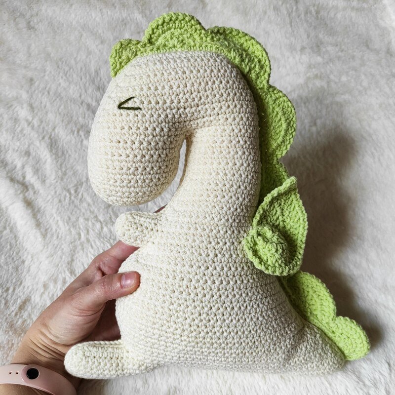 Off-white crocheted dinosaur plush with green spikes and wings, held by a hand on a white fluffy background.