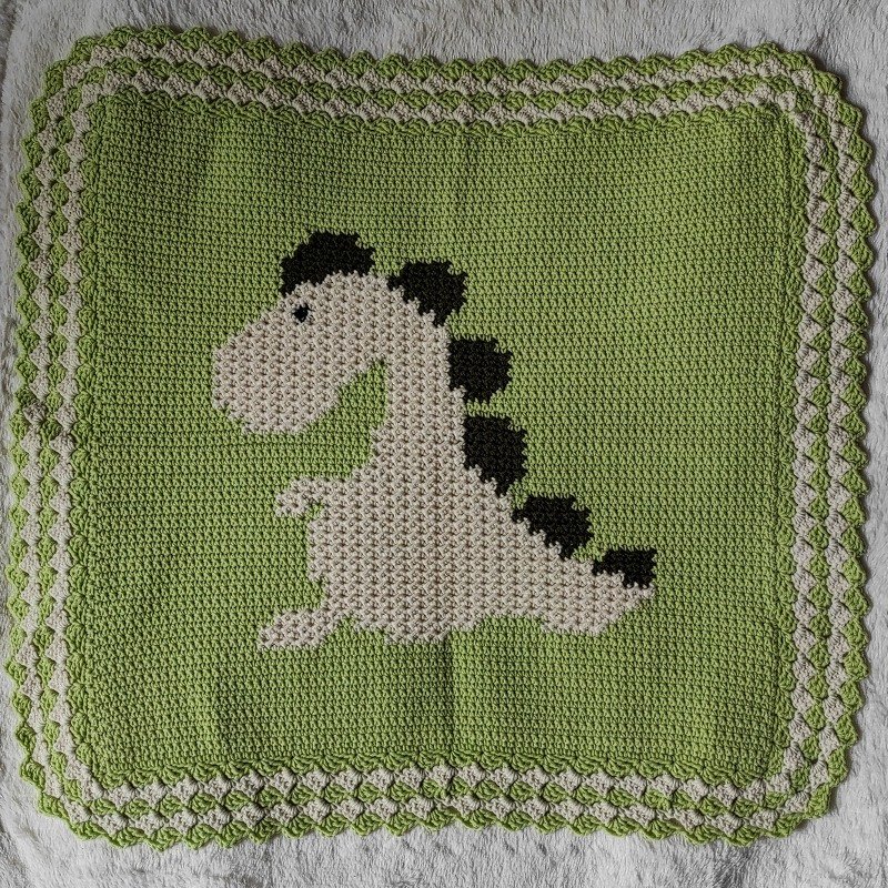 Light green crocheted baby blanket with white dinosaur motif and two-tone scalloped border.