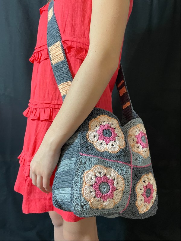 Gehäkelte Umhängetasche in Grau mit bunten Granny‑Squares und Blumenmotiven, über der Schulter getragen.