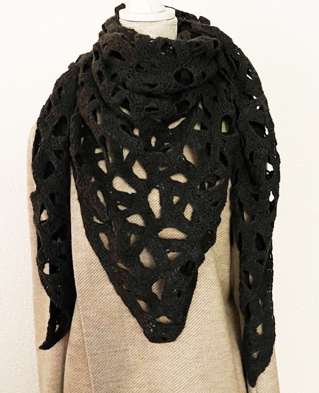 Black crocheted triangle shawl with openwork draped over a beige coat on a mannequin