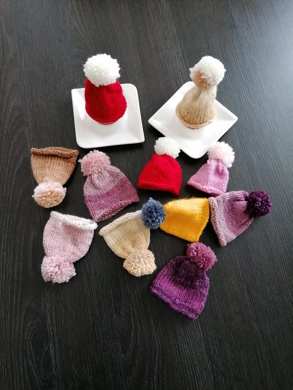 Mehrere bunte gestrickte Eierwärmer mit Pompons auf dunklem Holz, zwei auf kleinen weißen Tellern.