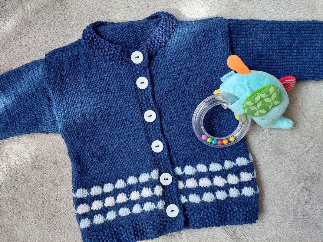 Dunkelblaue gestrickte Babyjacke mit weißen Knöpfen und weißem Noppenmuster, daneben ein weiches Spielzeug.
