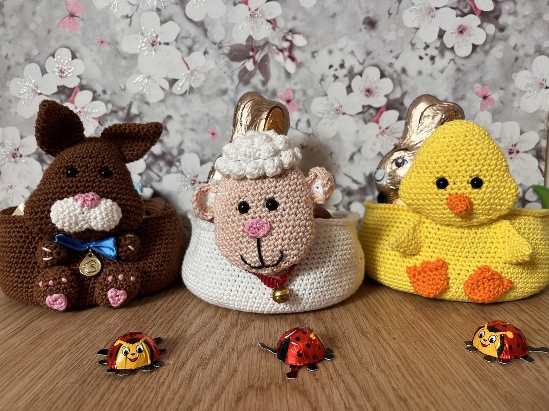 Drei gehäkelte Osterkörbchen (Hase, Lamm, Küken) auf Holzuntergrund vor blumigem Hintergrund, jeweils mit kleinen Schokoladenfiguren dekoriert.