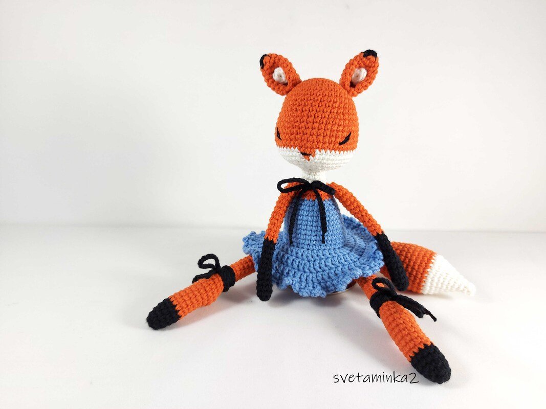 Crocheted orange fox amigurumi wearing a blue dress seated on a white background