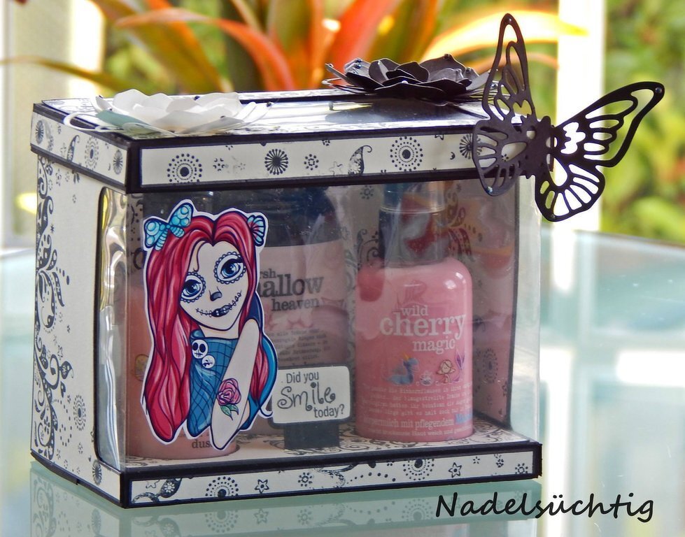 Clear acetate gift box with black patterned paper frame, filigree black butterfly on the corner, layered white paper flowers on the lid, colorful red-haired illustrated sticker on the front and a pink cosmetic bottle visible inside