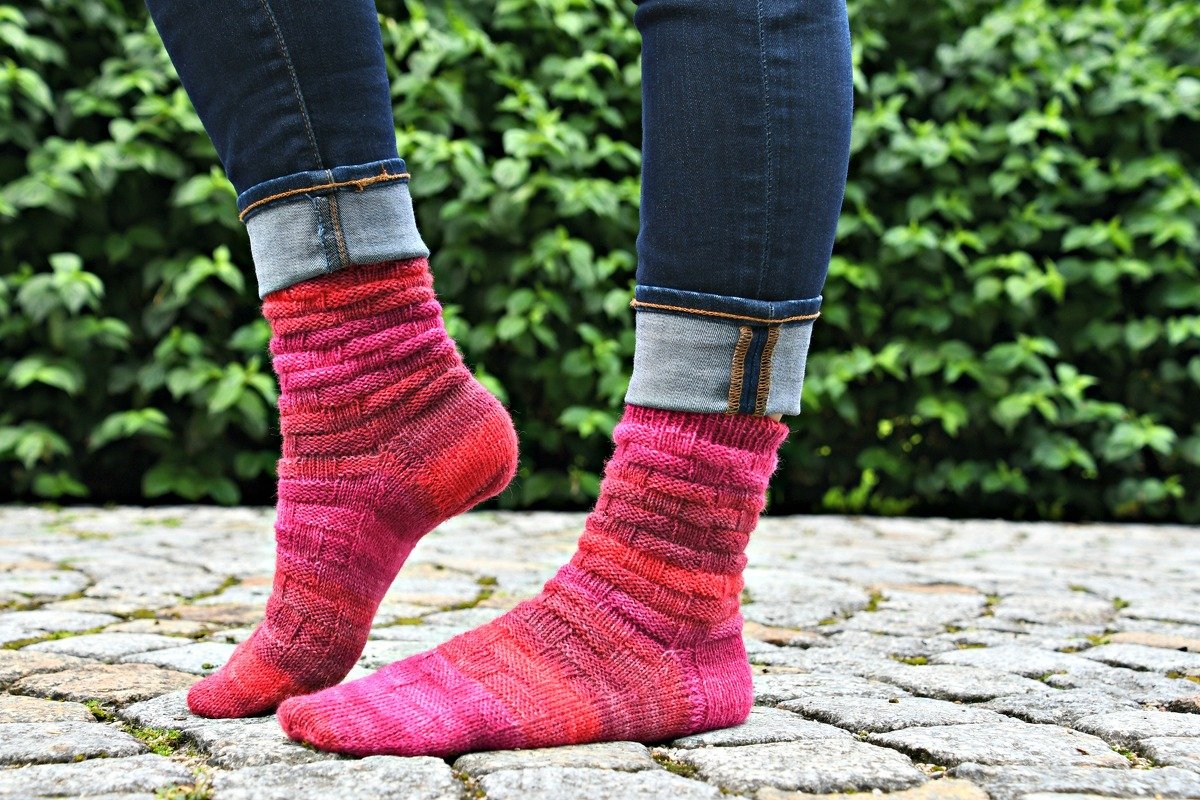 Nahaufnahme von hochgekrempelten Jeans und pink-rot gestrickten Damensocken mit Strukturmuster auf Kopfsteinpflaster vor grüner Hecke.