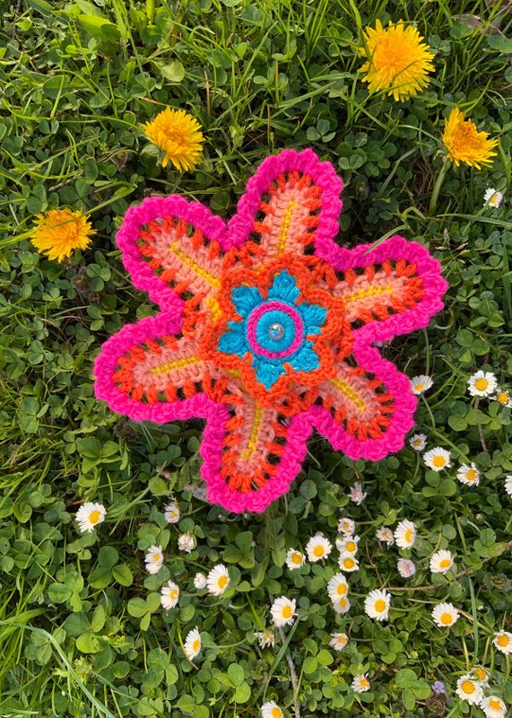 Bunte gehäkelte Blumenapplikation mit Zierknopf, liegend auf Gras mit Löwenzahn und Gänseblümchen.