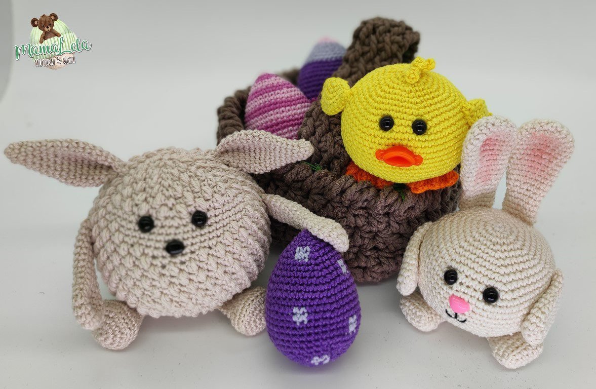 Zwei gehäkelte Amigurumi-Hasen, ein gelbes Küken im Häkelkörbchen und lila-rosa Häkeleier auf hellem Hintergrund.