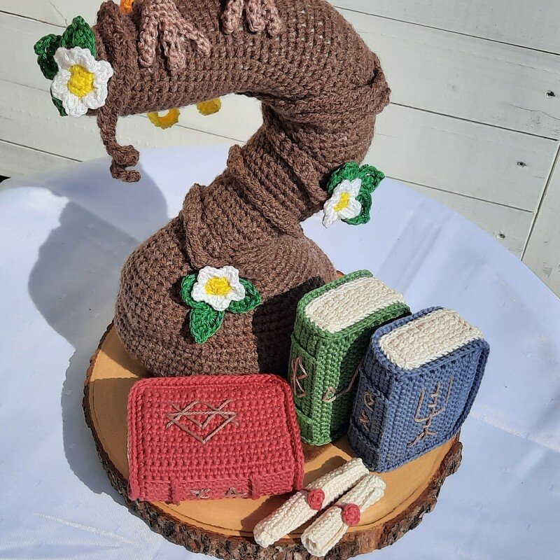 Gehäkelter brauner Vogel-Amigurumi mit weißen Blüten, drei kleinen gehäkelten Büchern und zwei Schriftrollen auf Holzscheibe