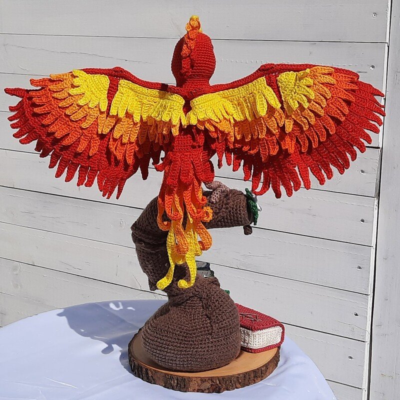 Gehäkelter Phönix in Rot, Orange und Gelb mit ausgebreiteten Flügeln auf rundem Holzsockel