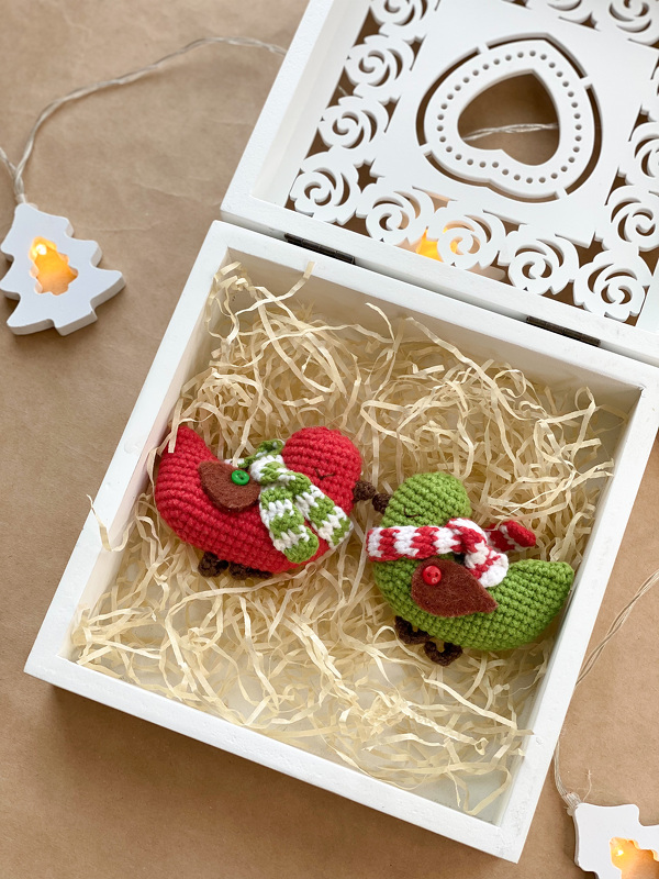 Two small crocheted bird amigurumi in red and green with striped scarves inside a white decorative box filled with shredded paper