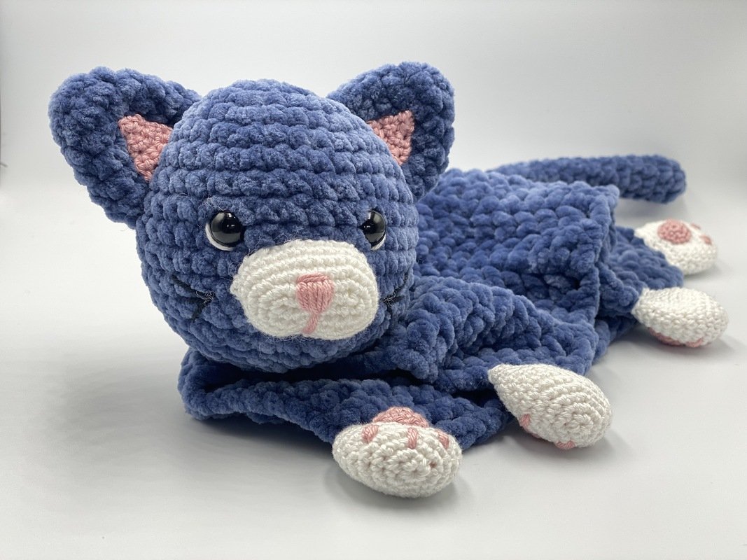Blue crocheted cat snuggle blanket with plush head, tail and white paws with pink pads on a white background