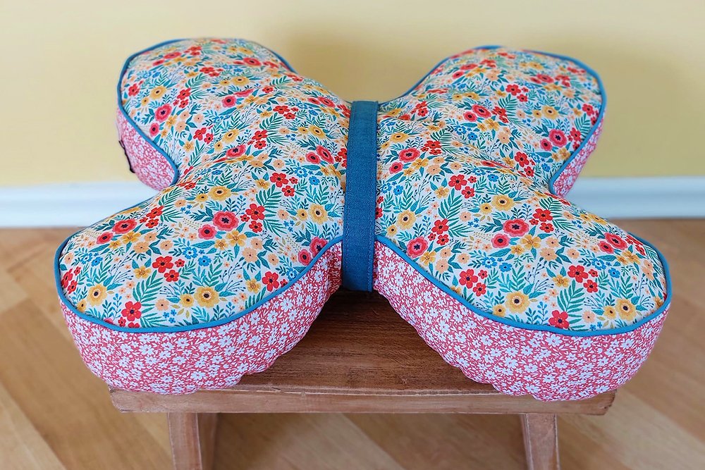 Schmetterlingskissen mit floralem Stoff, blauer Paspel und rotem Seitenteil auf Holzschemel