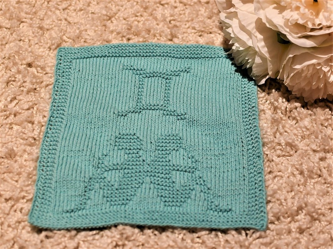 Turquoise knitted square showing an embossed Gemini zodiac glyph above two textured twin figures, with a garter stitch border